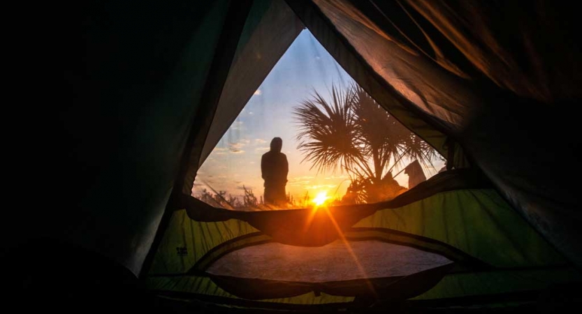From inside of a tent, the sun is rising on the horizon. A person stands outside the tent, looking towards the sunrise.
